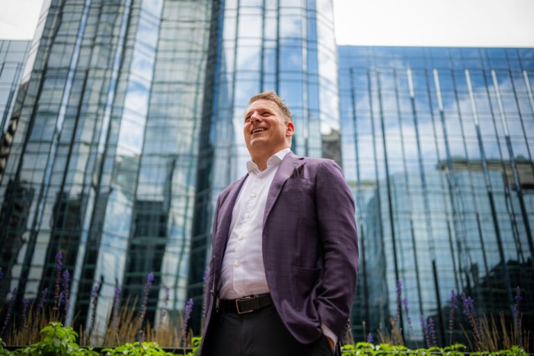Minto Group CEO Michael Waters in a purple blazer standing outside a modern glass building, smiling and looking slightly upward.