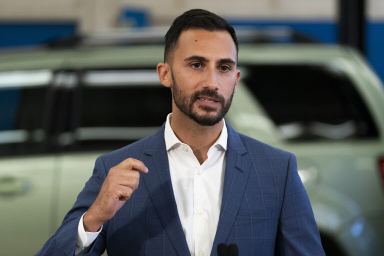 Stephen Lecce, who has dark brown hair and a short beard, speaks with his right arm partly raised for emphasis. He is wearing a blue blazer with a light tartan pattern, and a white shirt with no tie. There is a light green vehicle out of focus in the background.