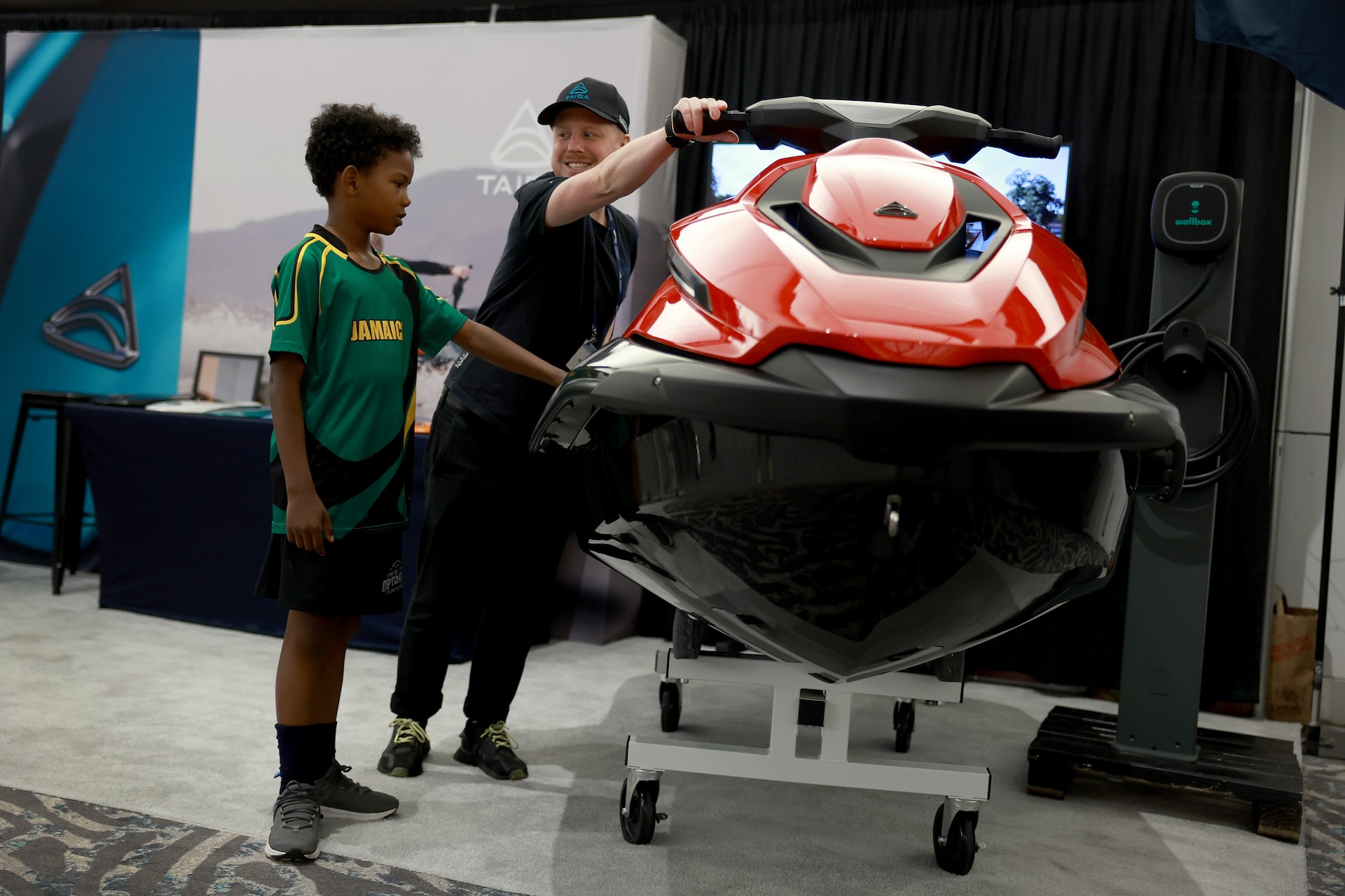 A person wearing a cap shows a boy in a Jamaica jersey a red watercraft on display in an indoor setting.