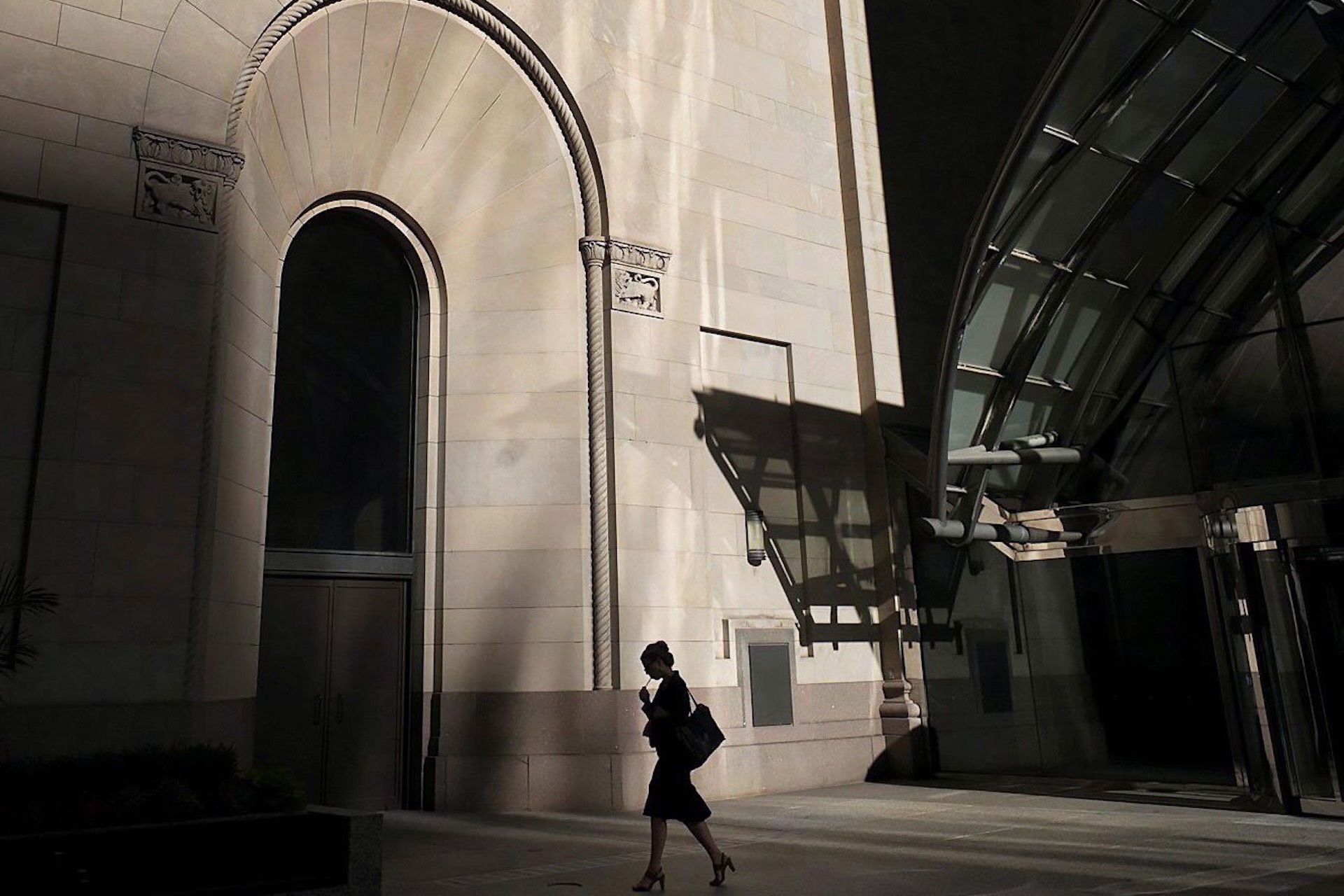 A silhouetted person walking down a street, in heels, wearing a purse, next to a tall building.