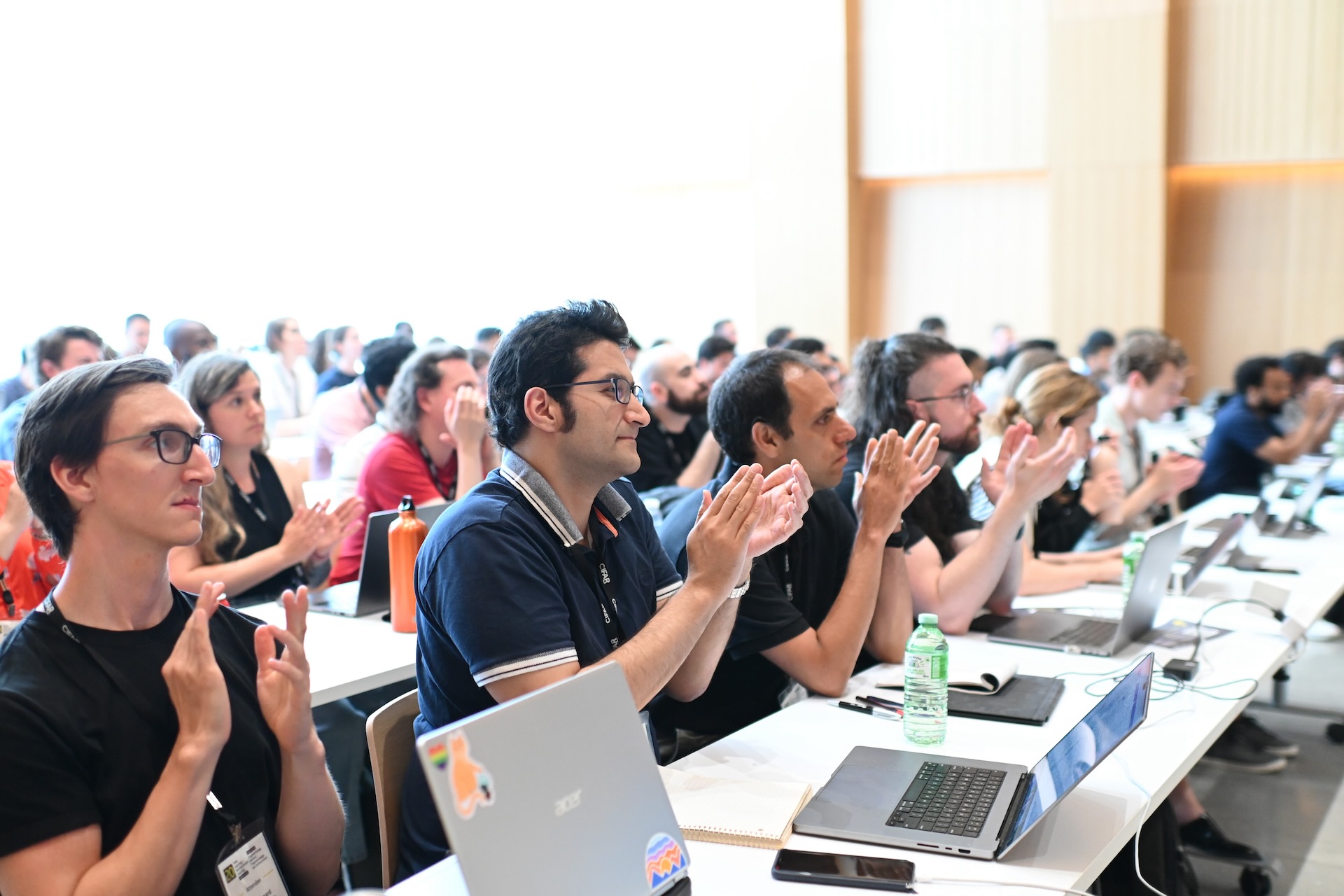 A group of students sitting at a desk with their laptops open in front of them. They are looking in front and clapping their hands.