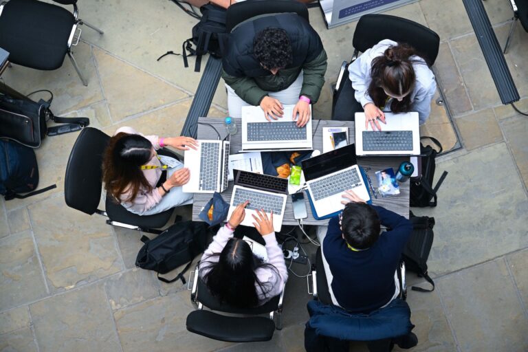 A shot from above of five people clustered around a table, all working on near-identical laptop computers. Their computer bags lie on the floor and some are wearing yellow lanyards.