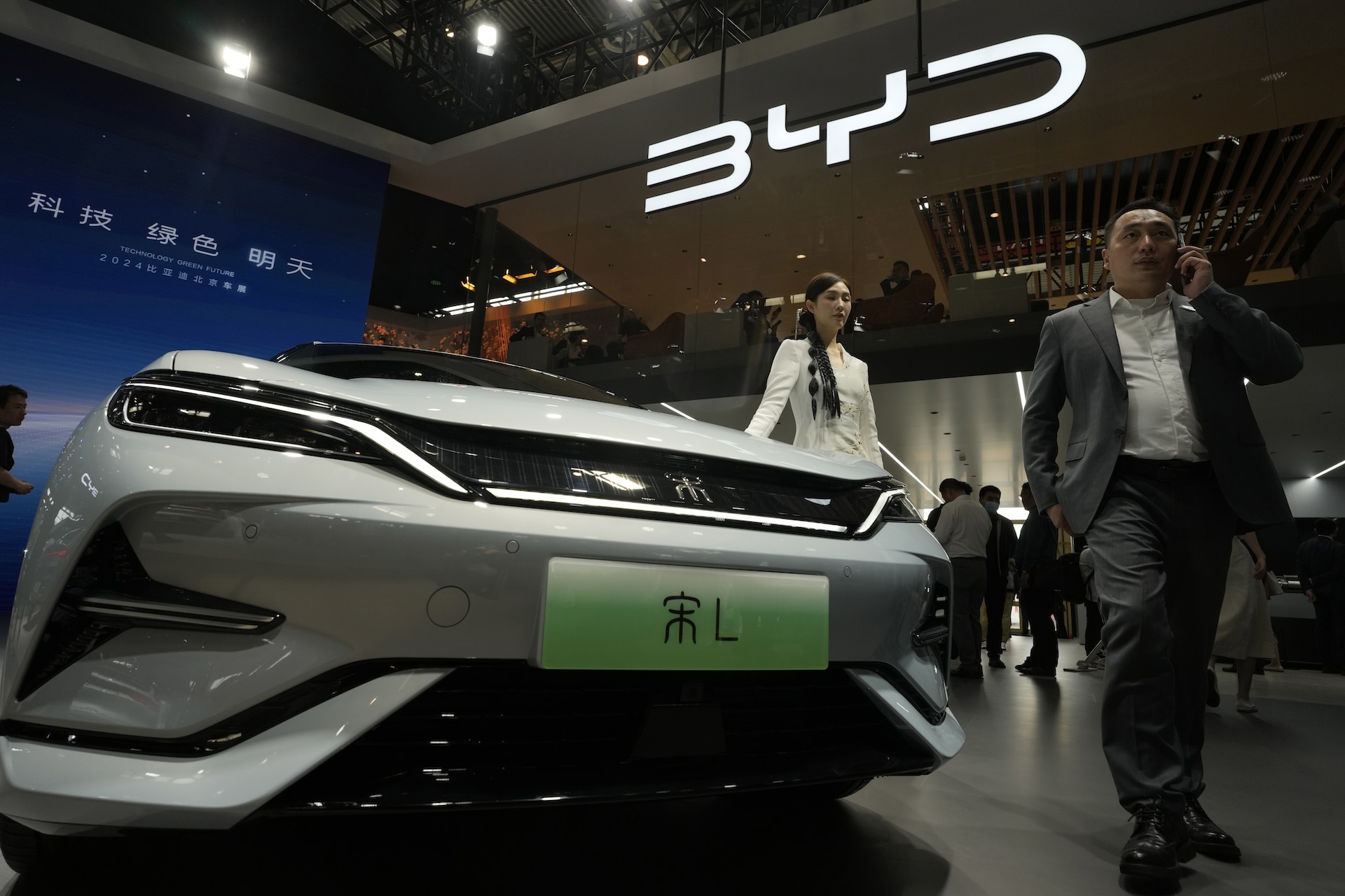 A white BYD electric car is showcased at an indoor auto exhibit. A man in a suit and a woman in a white dress are walking nearby. The BYD logo is prominently displayed above.