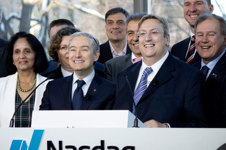 Mark Barrenechea, wearing a dark suit, frameless glasses and a striped tie, stands at a lectern with François-Philippe Champagne to his right and a cluster of others behind him. There are tall windows in the background.