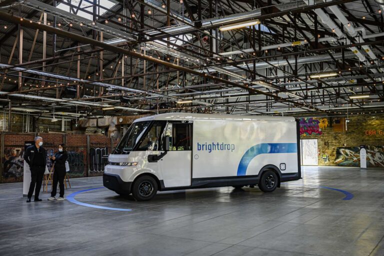 A Brightdrop electric delivery vehicle at an indoor event. Two people in black, wearing masks, are standing on the left.