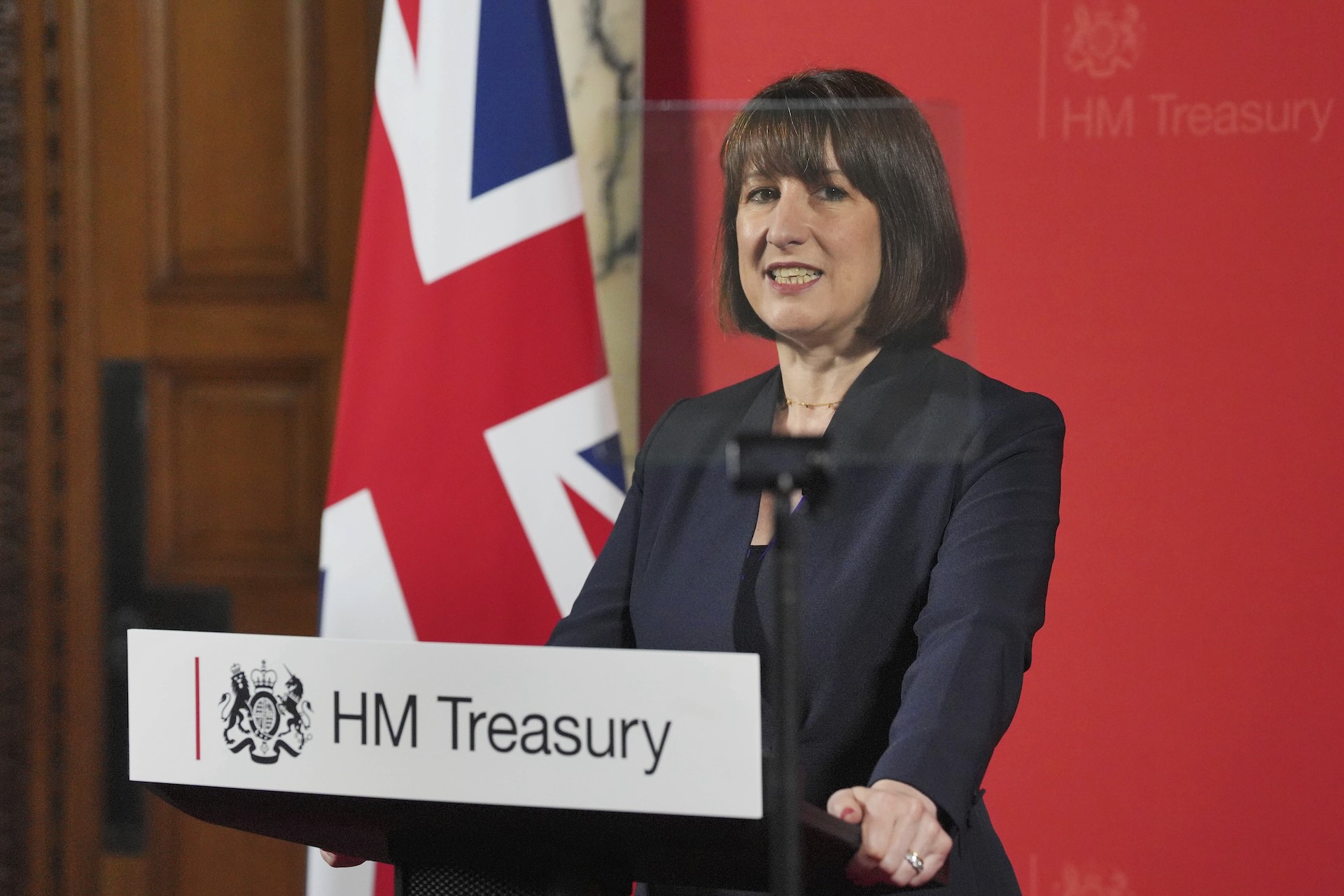 U.K. Chancellor Rachel Reeves standing at a podium wearing a black suit. There’s a British flag and a red HM Treasury backdrop behind her.
