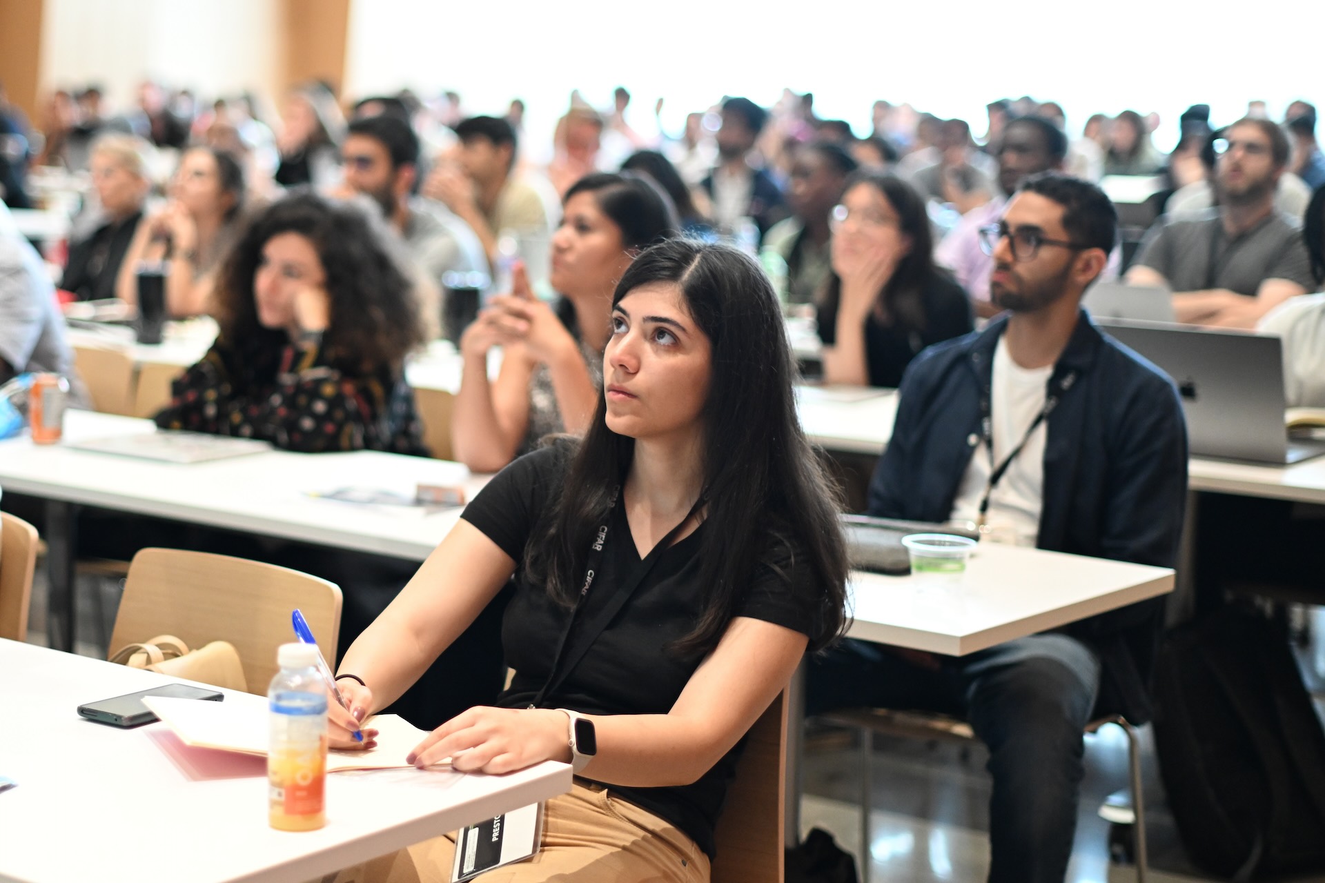 A group of people sitting at tables in a conference room, attentively listening and taking notes during a presentation.