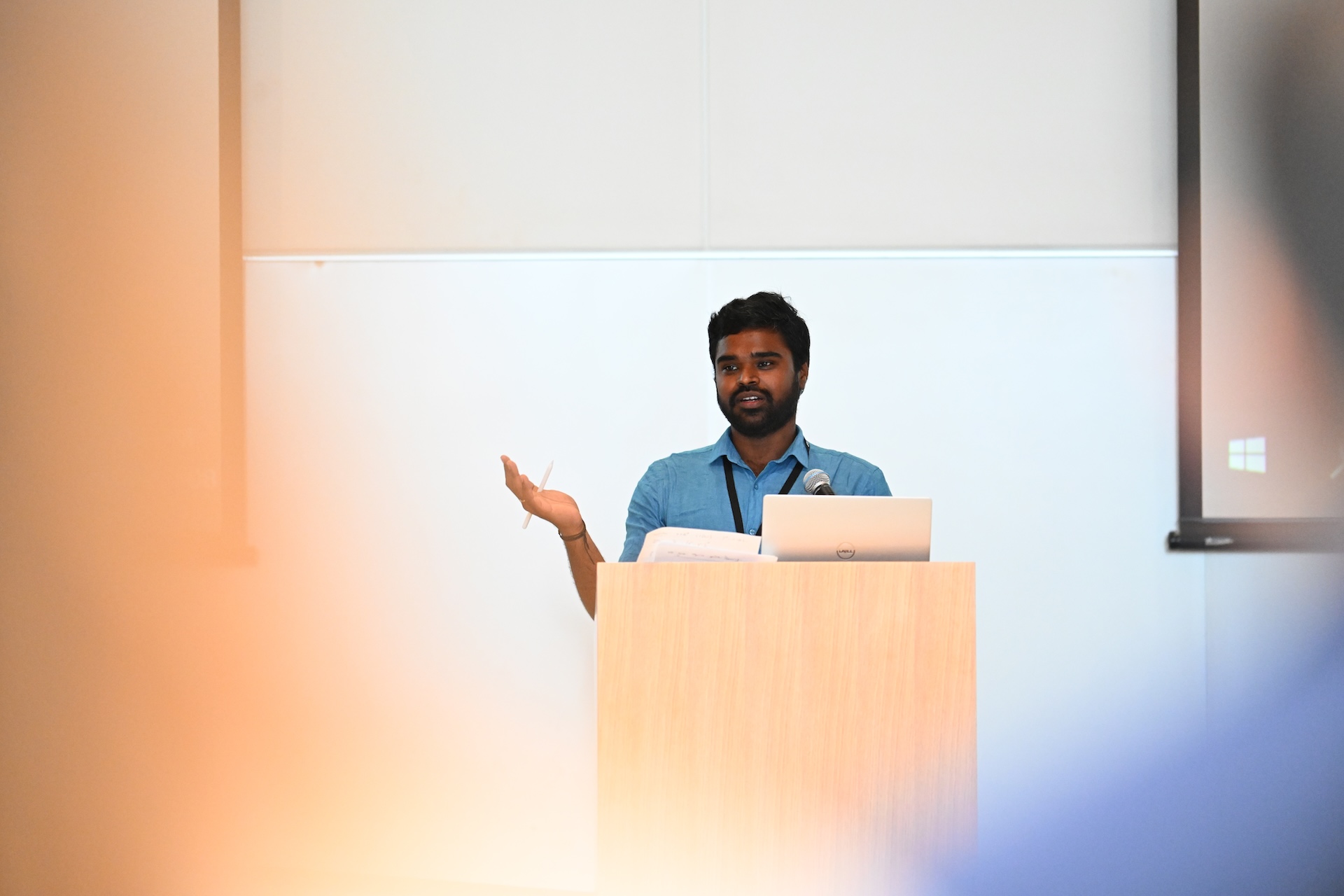Sarath Chandar, wearing a blue button-down shirt and holding a pen, gives a lecture on a podium. A laptop rests on the podium and a projector screen behind him displays a Microsoft Windows logo.