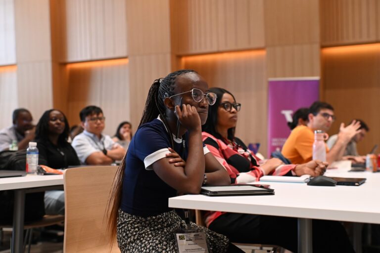 A diverse group of people attentively listen to a presentation in a conference room. Some take notes while others observe the speaker off-camera.