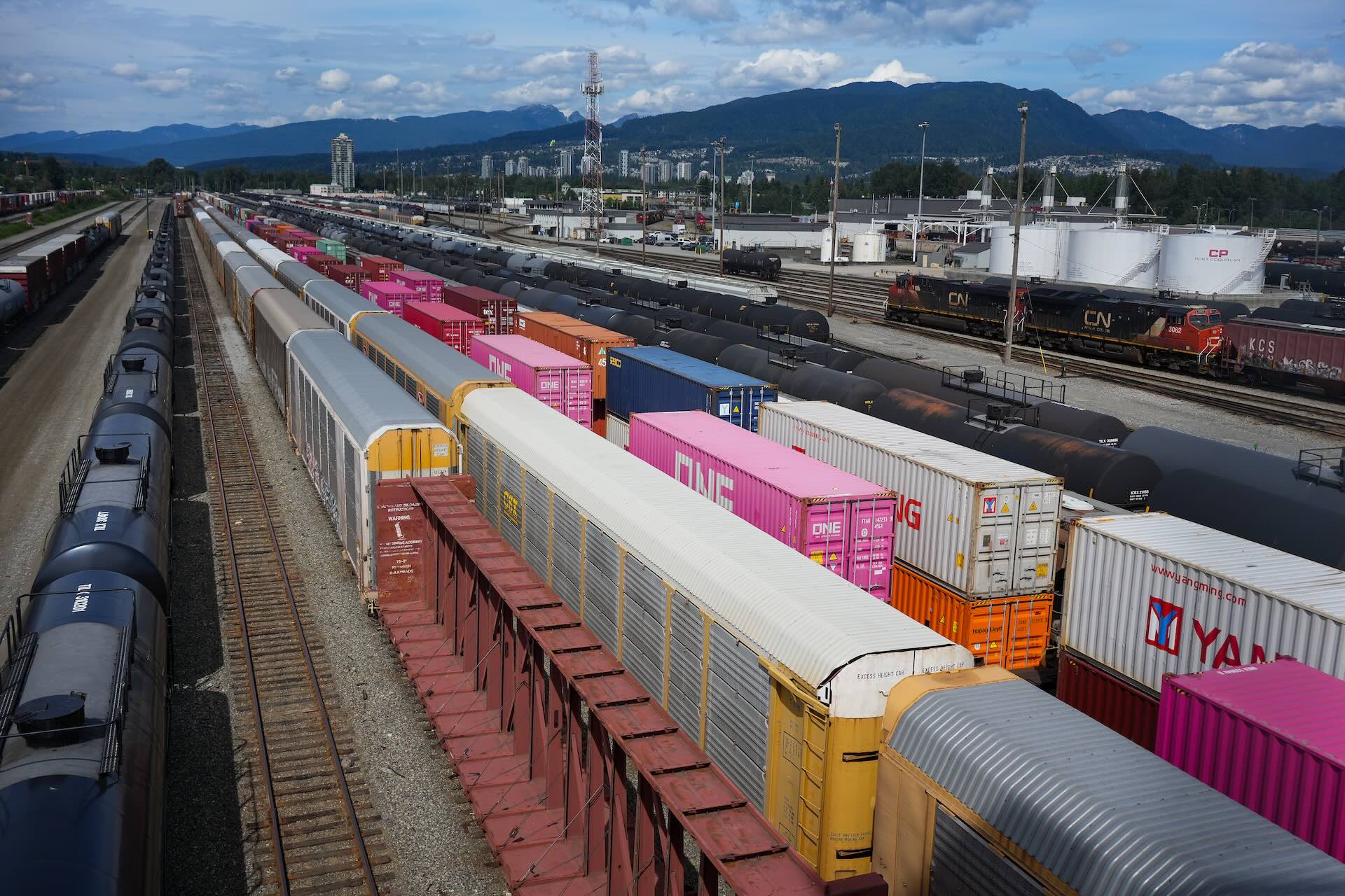 An aerial view of a freight train rail yard with multiple colourful shipping containers and tank cars on tracks. Industrial facilities and mountains are visible in the background under a cloudy sky.