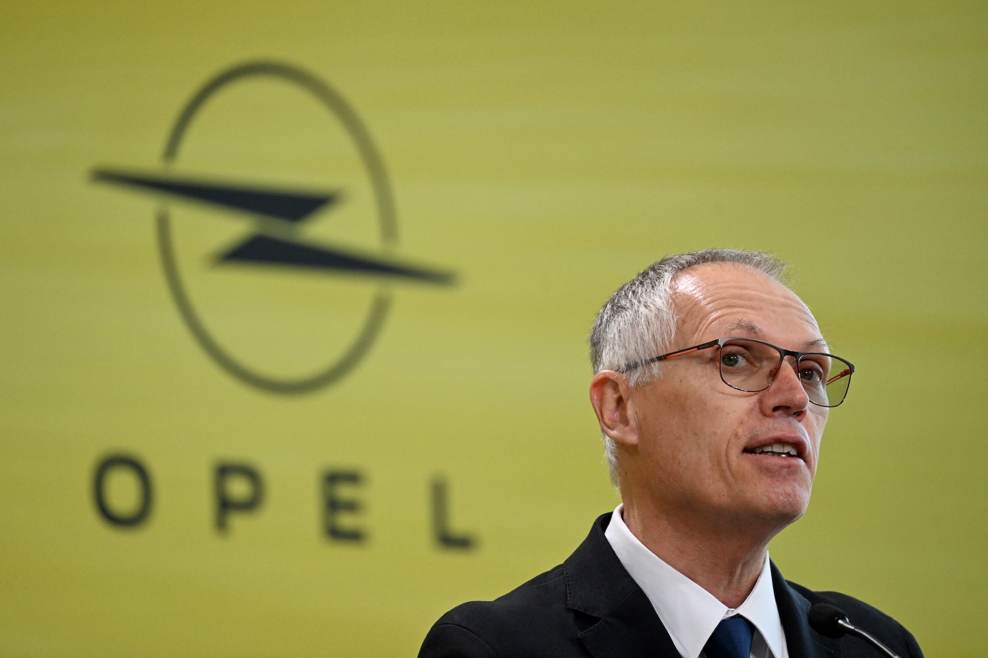 Stellantis CEO Carlos Tavares wearing a suit and giving a speech, with a yellow background that reads “Opel” and displays a logo with a lightning bolt inside a circle.