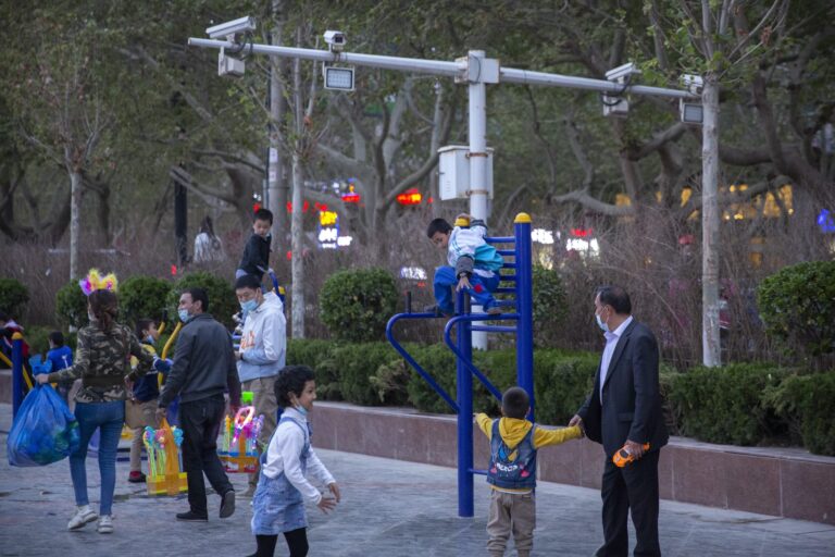Children climb on playground equipment while adults, some wearing masks, supervise in a public square lined with trees and shrubs. Surveillance cameras can be seen on a gantry above the playground equipment.