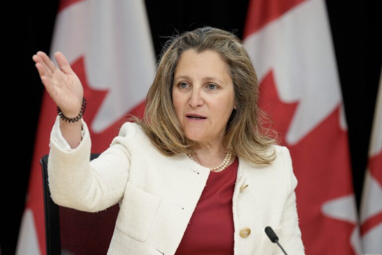 Finance Minister Chrystia Freeland, wearing a red shirt and white blazer jacket, gestures with her right arm at a press conference with Canadian flags in the background.