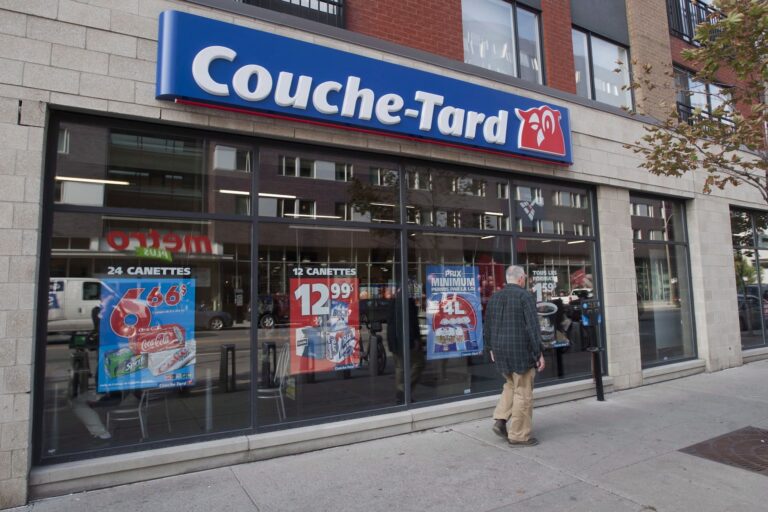 A man in a dark plaid shirt walks past a Couche-Tard convenience store. There are posters in the windows advertising pop and beer.