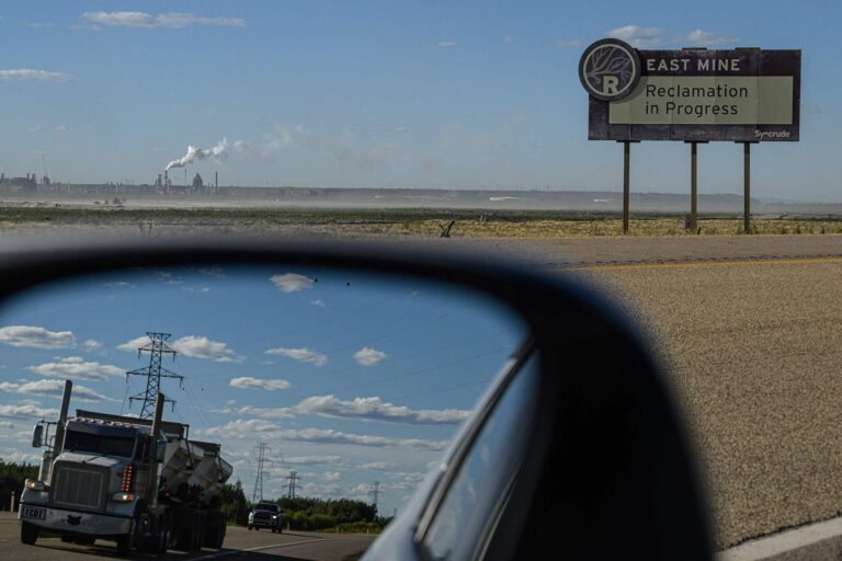 A view from the roadside of an oil sands plant, with a sign that reads "East Mine Reclamation Project" in middle distance. Also in the frame is the car's sideview mirror, which shows an approaching dump truck.