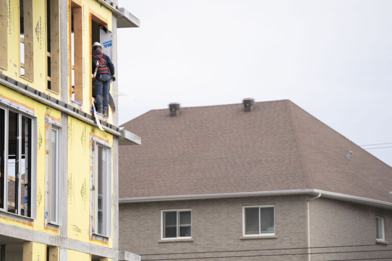 A worker stands with his back to the camera in the window opening of a partially constructed building. He's wearing a safety harness and white hardhat, and there's fully built house in the background.
