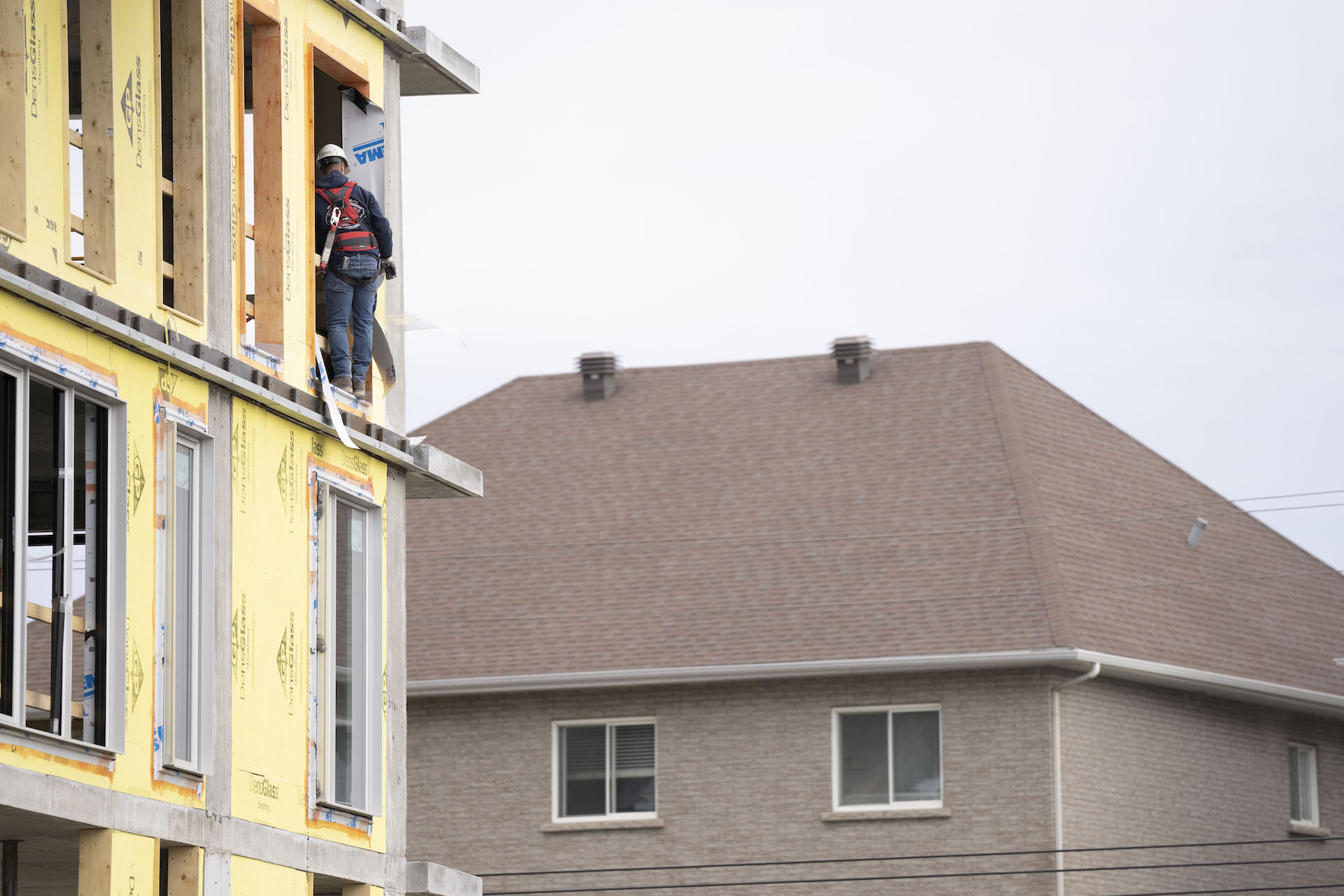 A worker stands with his back to the camera in the window opening of a partially constructed building. He's wearing a safety harness and white hardhat, and there's fully built house in the background.
