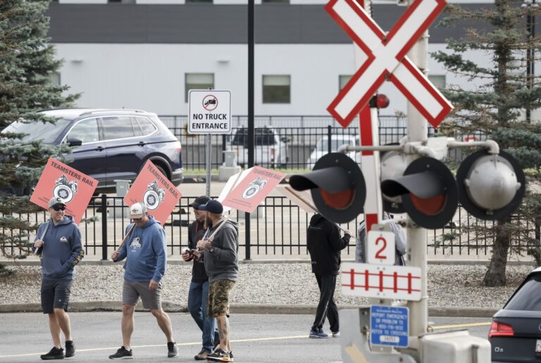 Unionized railway workers wearing hoodies, shorts and ballcaps walk around a parking lot carrying red-and-black Teamsters signs. There is a railway crossing signal poll, with lights and a traffic arm, in the foreground.