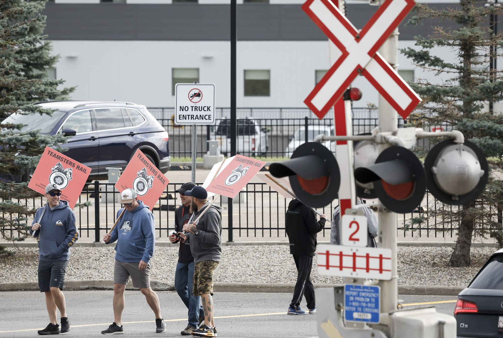 Unionized railway workers wearing hoodies, shorts and ballcaps walk around a parking lot carrying red-and-black Teamsters signs. There is a railway crossing signal poll, with lights and a traffic arm, in the foreground.