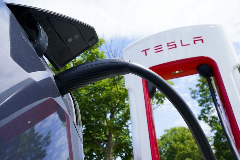 A close-up of a Tesla electric car charging at a supercharger station on a sunny day, with trees and a blue sky in the background.