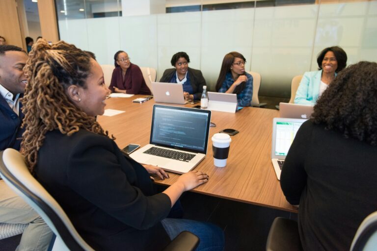A group of people sitting around a conference table engaged in a meeting. Some have laptops open and one person has a coffee cup.