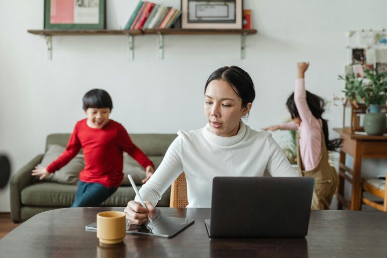 A woman sitting at a desk holds a stylus pen over a tablet device. A laptop screen is open in front of them and there are two children running and playing in the background.