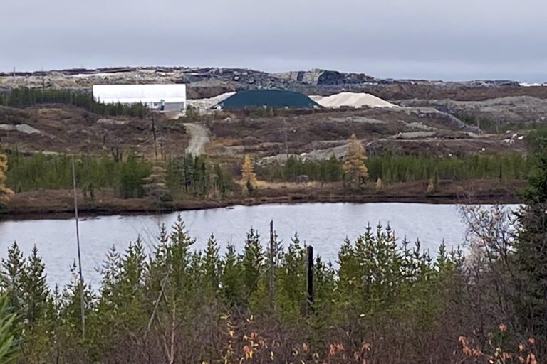A view of a mine site in low, rocky hills, taken across a pond, with scrub brush in the foreground. A large metal-clad building and piles of slate- and cream-coloured ore are visible in middle distance.