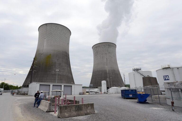 Two concrete cooling towers of a nuclear power plant loom into a cloudy sky. There is steam coming from the tower on the left.