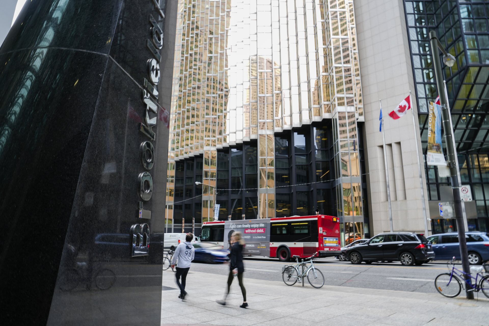 People walking past the building displaying Brookfield Place signage with reflections of skyscrapers, a red bus, and Canadian flags visible on a busy street.
