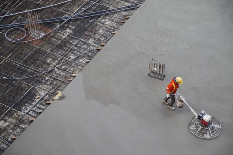 An overhead shot of construction worker in an orange reflector jacket and yellow hardhat power-trowelling a concrete surface.