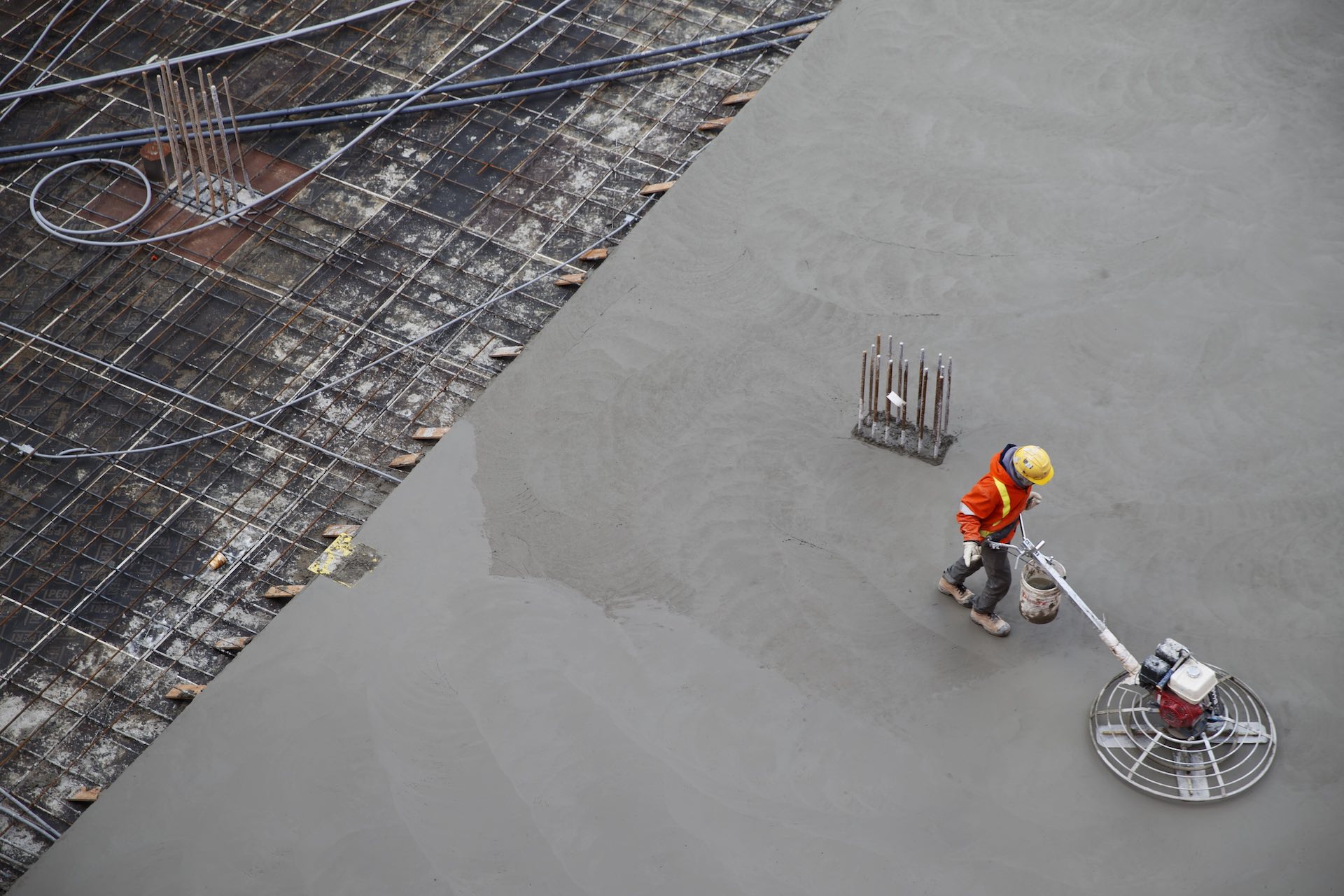 An overhead shot of construction worker in an orange reflector jacket and yellow hardhat power-trowelling a concrete surface.