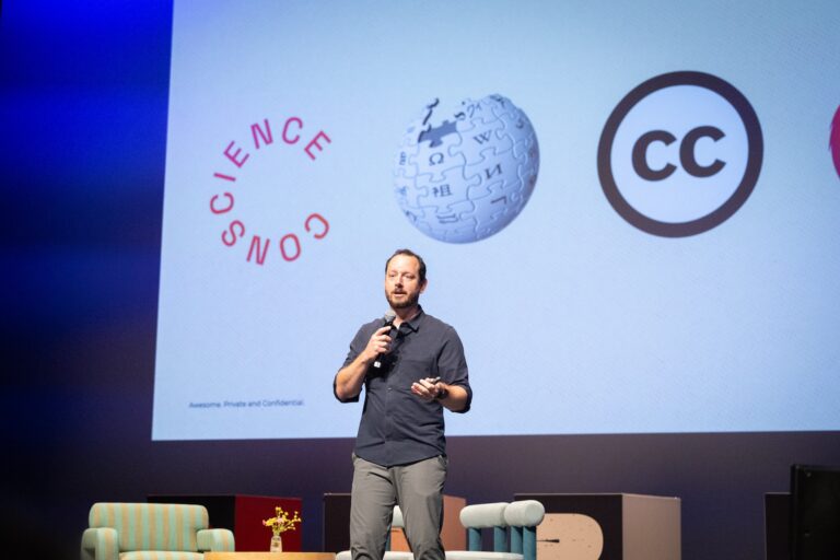 Ryan Merkley stands with a microphone in front of a large screen displaying logos, including those of Conscience and Wikipedia. He has dark hair and a short beard, and is wearing a slate-black, button-up shirt with the sleeves partly rolled up.