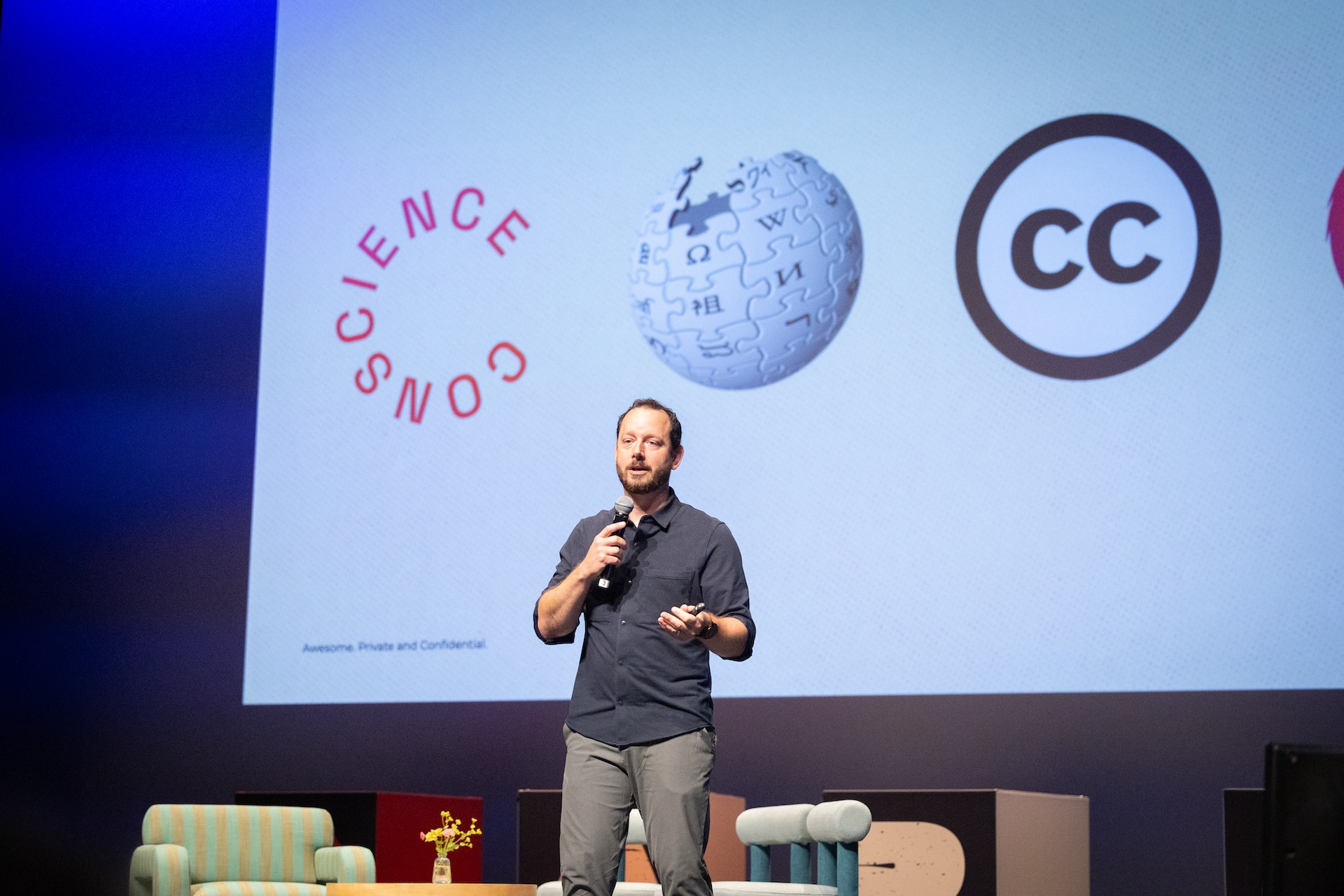 Ryan Merkley stands with a microphone in front of a large screen displaying logos, including those of Conscience and Wikipedia. He has dark hair and a short beard, and is wearing a slate-black, button-up shirt with the sleeves partly rolled up.