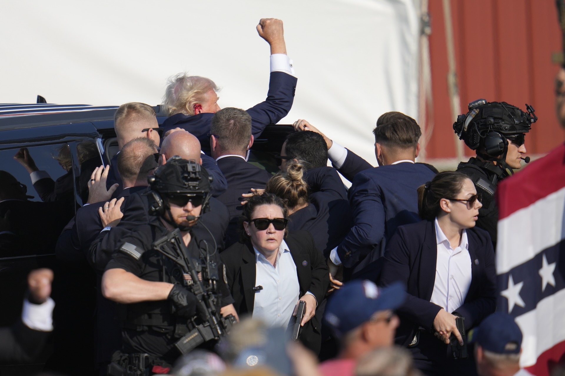 Donald Trump climbs into a car with blood visible on his ear and his arm raised. He is surrounded by security staff, including two men wearing black high-tech helmets and carrying automatic weapons.