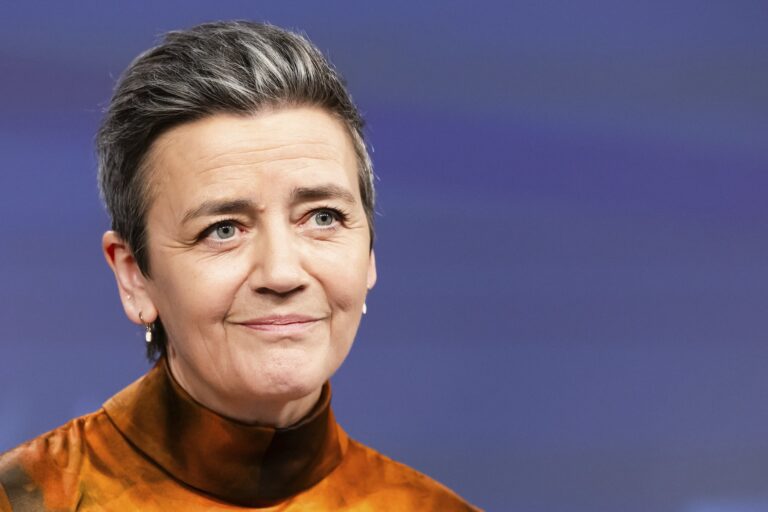 EU Commission vice president Margrethe Vestage, wearing a high-necked brown and orange top, gazes slightly to the side against a blue background.