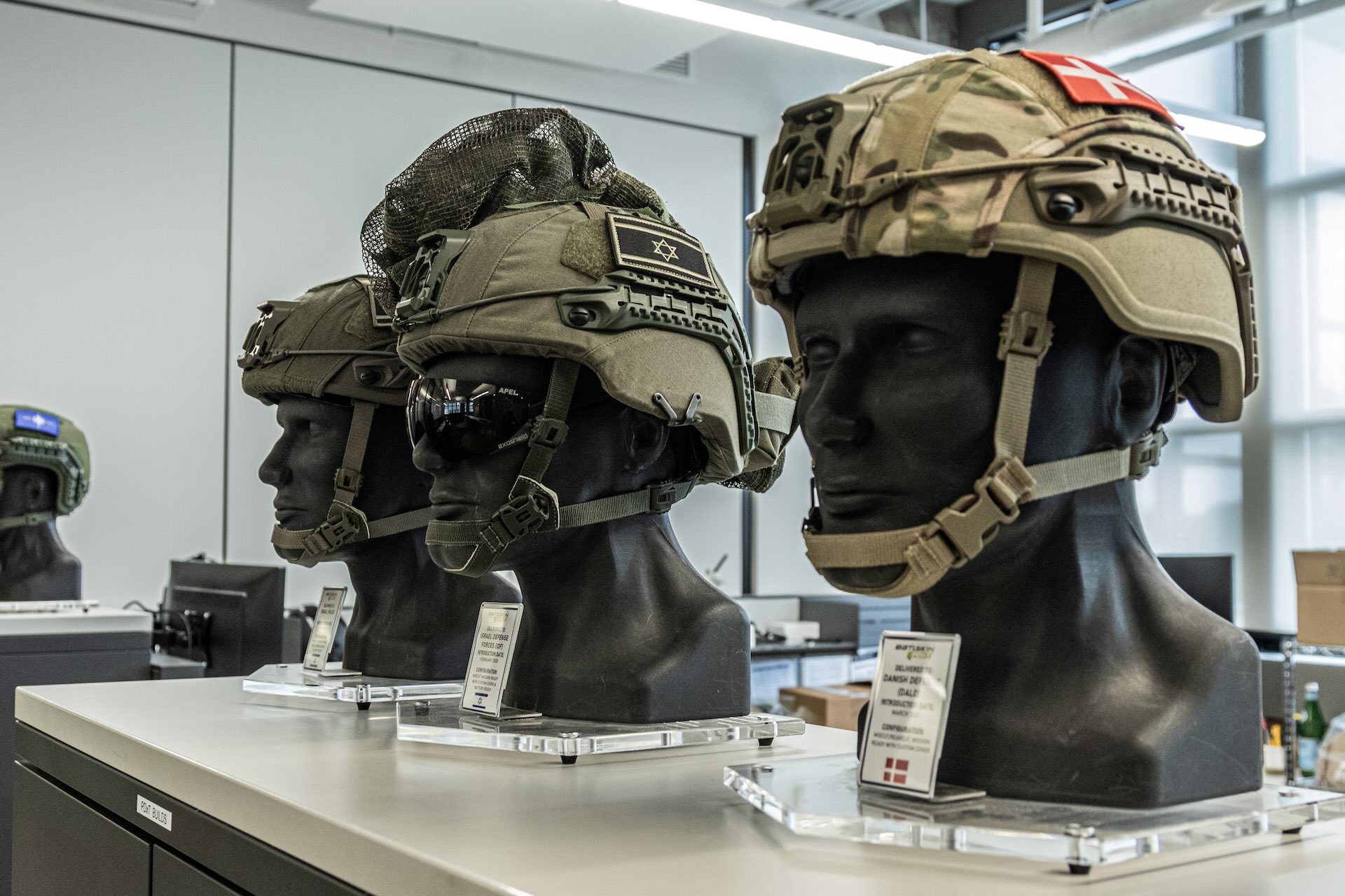 A close-up image of three mannequin heads wearing Galvion helmets at the company’s headquarters. Israeli and Danish flags are visible on two helmets. A fourth helmet in the background has a NATO flag.