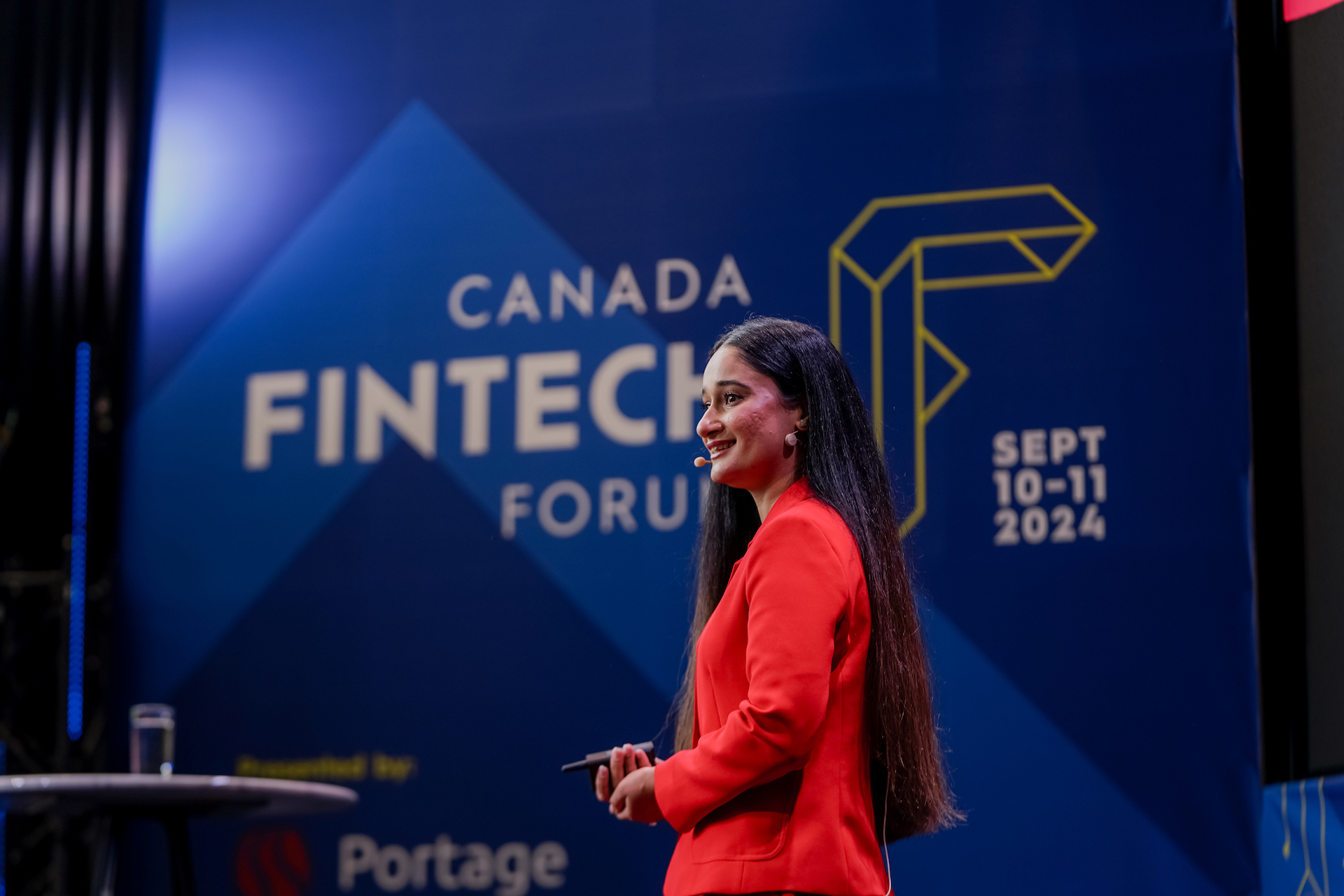 Jeet Bhatti stands smiling before a blue backdrop that bears the Canada Fintech Forum logo. She's wearing a bright, red blazer and has a headset microphone near her right cheek.