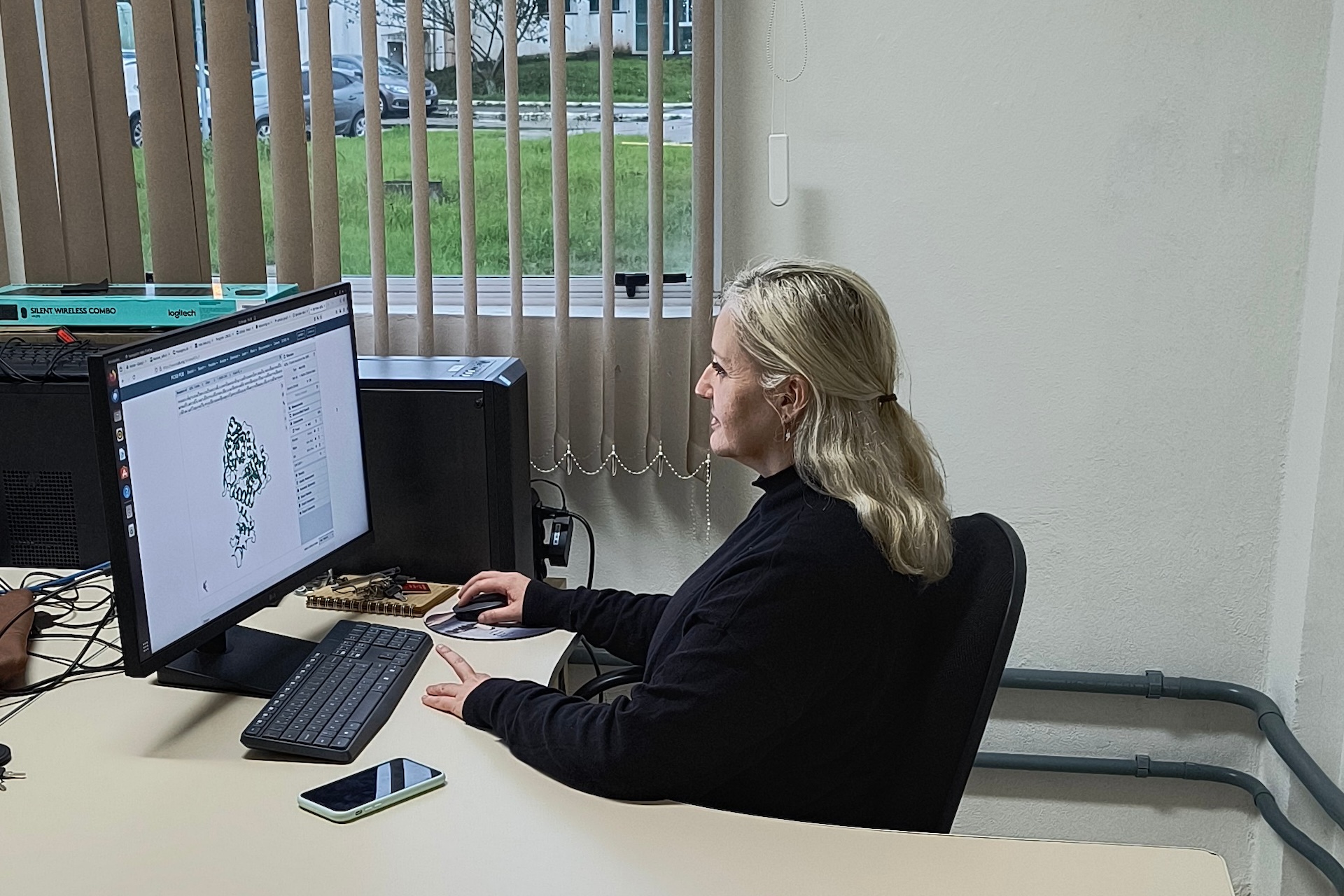 Machado is seated before a computer monitor at an office work table. Parked cars and tall grass are visible through a window in the background.