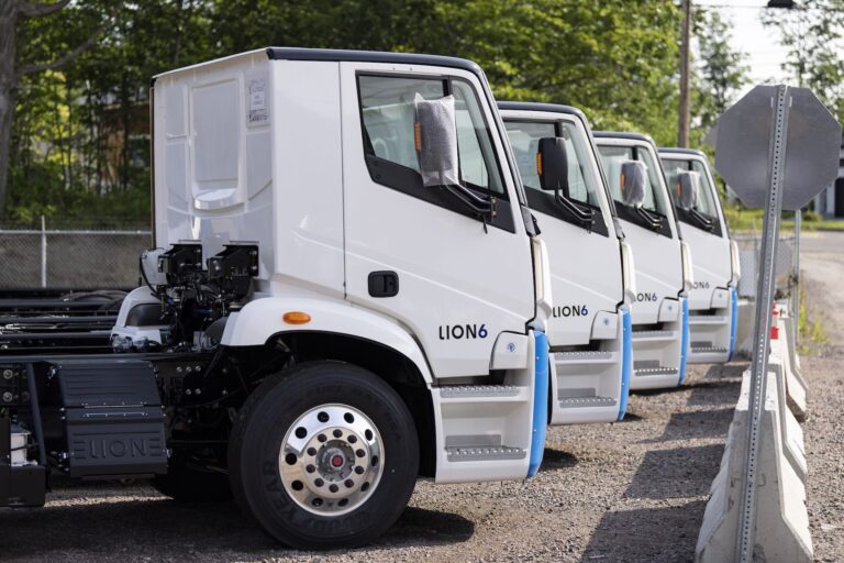 A side view of four new, white trucks parked in a row. The trucks are flat-faced and have the model name "Lion 6" stencilled on their doors.