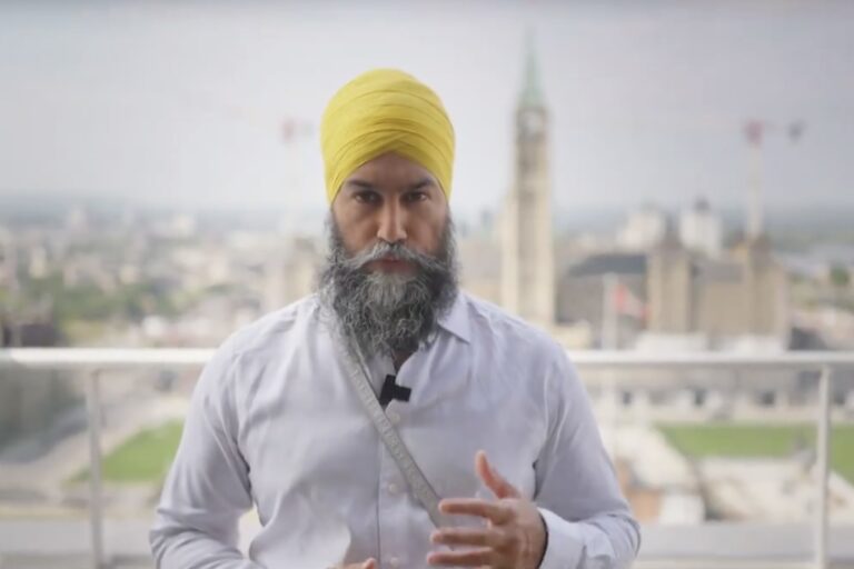 NDP Leader Jagmeet Singh, wearing a white button-up shirt and yellow turban, looks directly into the camera with his left hand slightly raised. In the background is the Peace Tower of the Parliament Buildings in Ottawa.