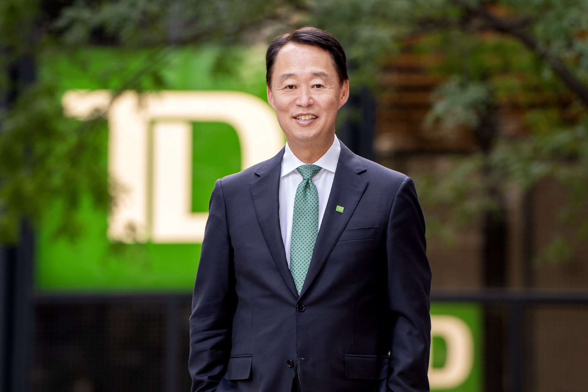 Raymond Chun wearing a navy suit and a green tie, with trees and the TD logo visible in the background.