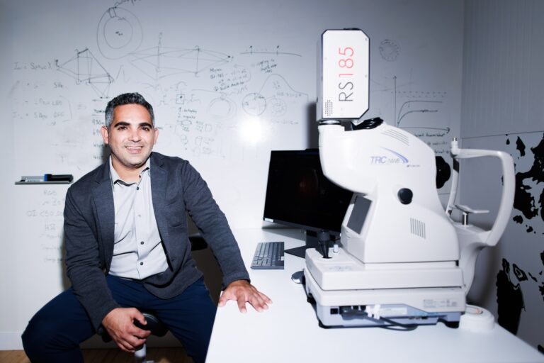 A man sits beside a large, white medical imaging device labeled "RS 185" in a lab with detailed diagrams on a whiteboard behind him. A computer monitor and other lab equipment are visible.