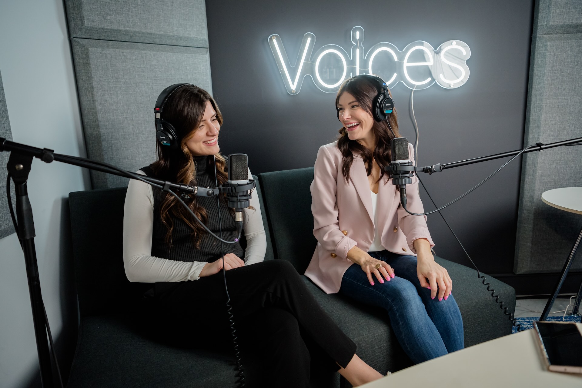 Two women, seated on a couch and wearing headphones, are engaged in a lively conversation in front of microphones, with a neon "Voices" sign glowing in the background.