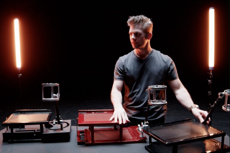 ohn Q. Kubin, one of the top featured talents on Voices.com, is standing in front of various equipment, illuminated by two vertical light fixtures in a dark room, adjusting a piece of machinery with his right hand.