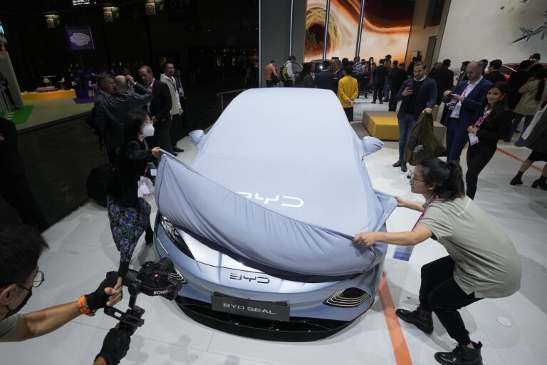 Attendants at a car show peel back a grey cover bearing the BYD logo from the front of a sleek, silver-blue vehicle. Only the nose of the car, which also has the BYD logo is visible.