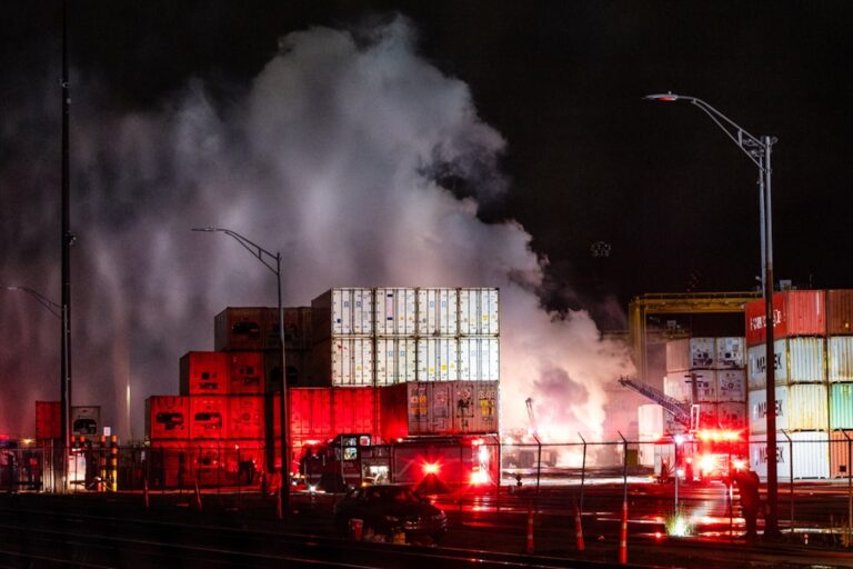 A night-time shot of white smoke rising over shipping container stacks, illuminated by white and red lights from emergency vehicles. The boom of a firetruck is visible on the right side of the frame.