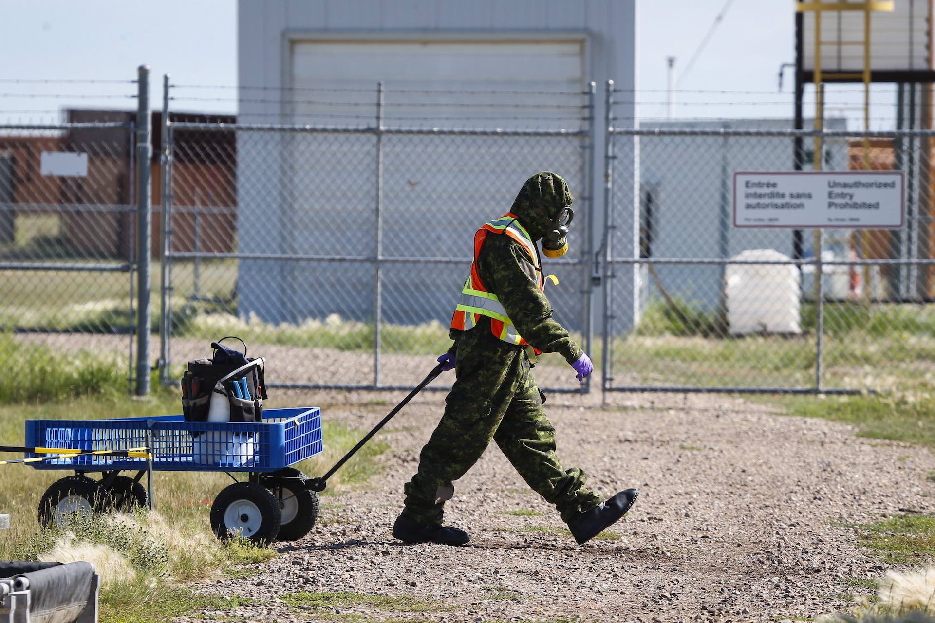 A soldiers wearing a camouflage hazmat suit, gas mask and orange reflector vest pulls a small wagon containing a white jug and tools across a road, with a chainlink fence and metal buildings in the background. On the fence is a sign reading "Unauthorized Entry Prohibited."