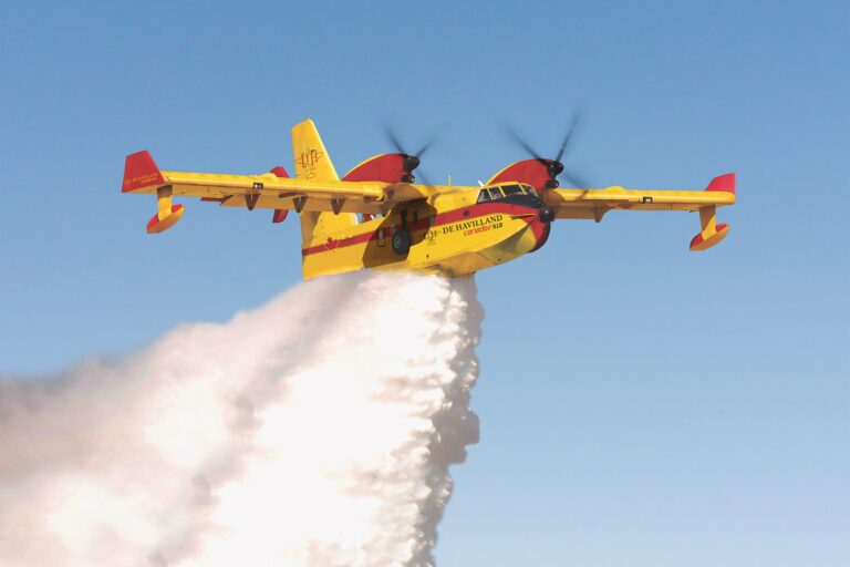 A large, yellow and red aircraft drops a white plume of water against a backdrop of clear blue sky. The plane has two engines mounted on the tops of its wings near the cabin, and small floats on the ends of the wings.