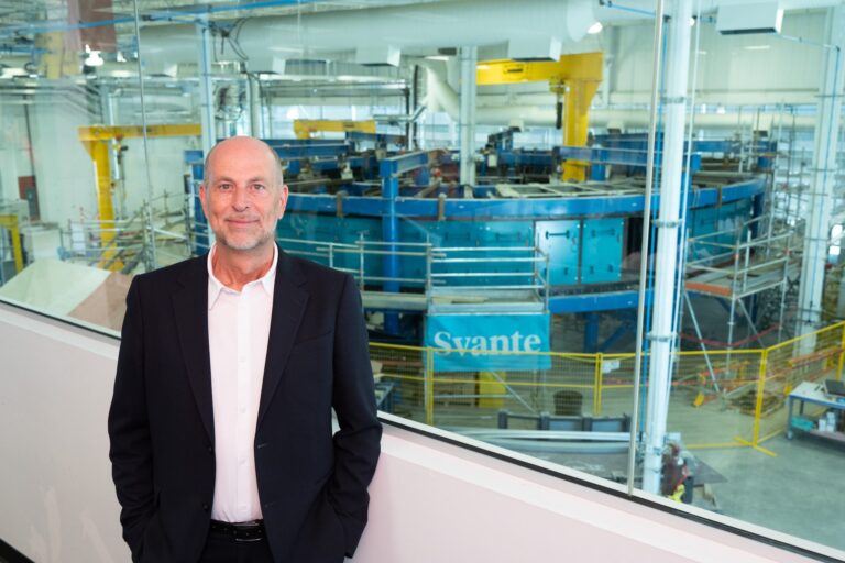 Svante CEO Claude Letourneau stands in a black suit and white open-necked shirt before a bank of windows overlooking the floor of a large industrial building. In the background, occupying much of the floor, is a large, circular piece of equipment painted blue and yellow. It's about the height of two-storey home.