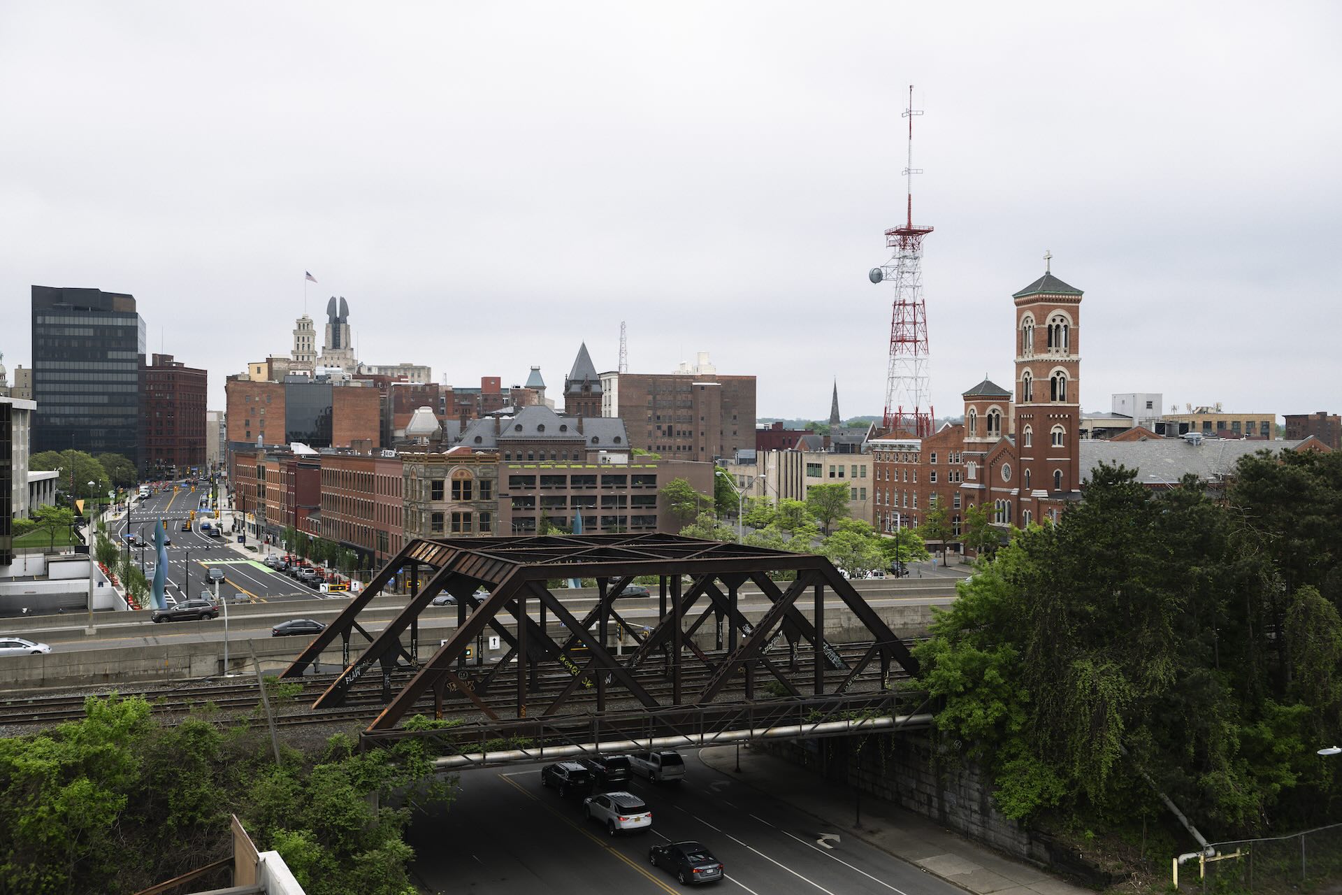 Cityscape featuring a mix of modern and historic buildings, a red and white communication tower, and a metal bridge over a highway with cars.